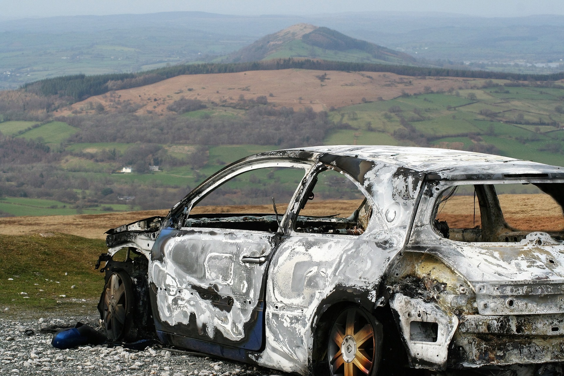 A burnt car, not from the story, stands in a field on a hillside with larger hills and mountains in the background.