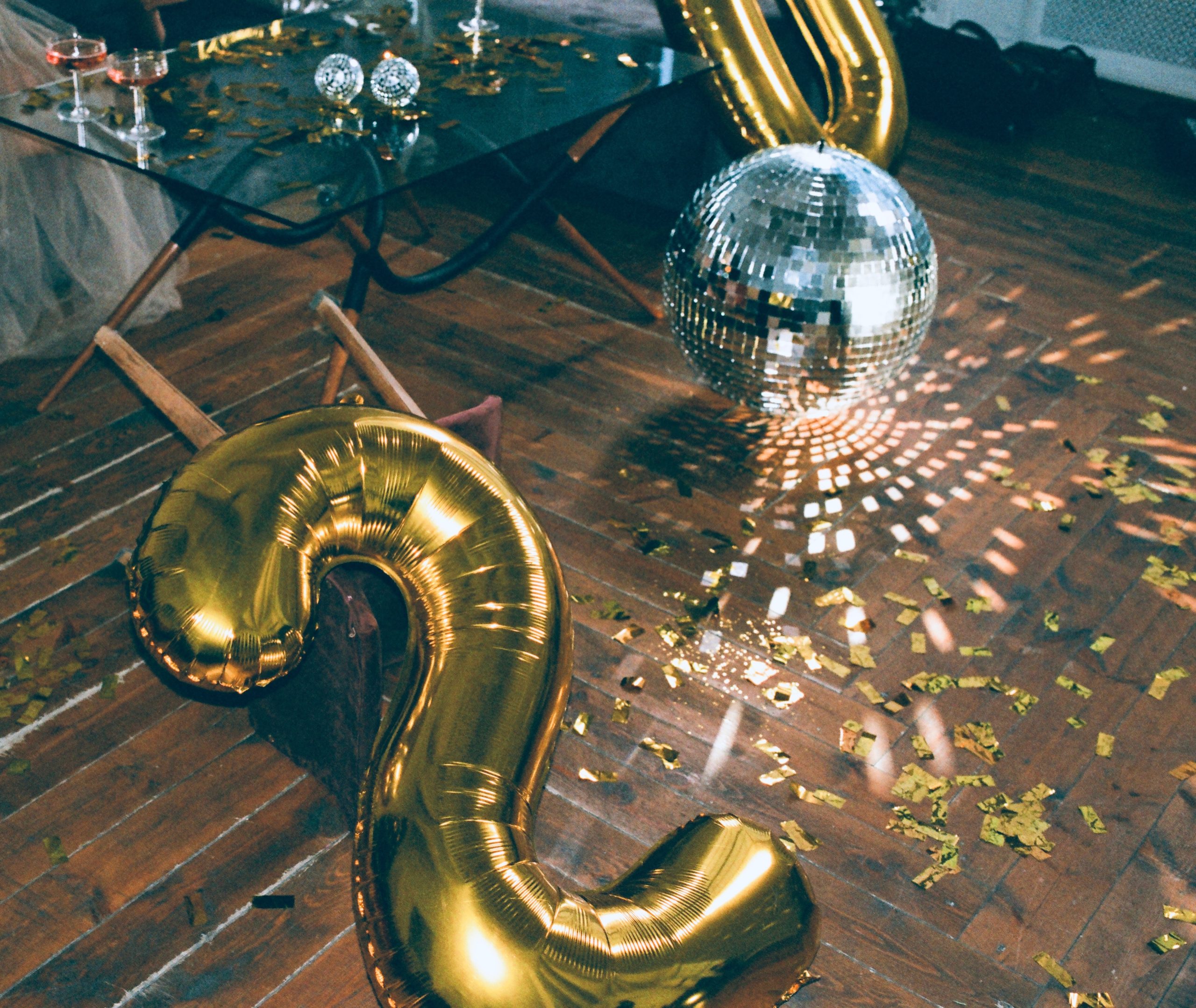 A gold balloon in the shape of the letter S lays on a wood floor near a disco ball and another gold ballon in a scene from party.