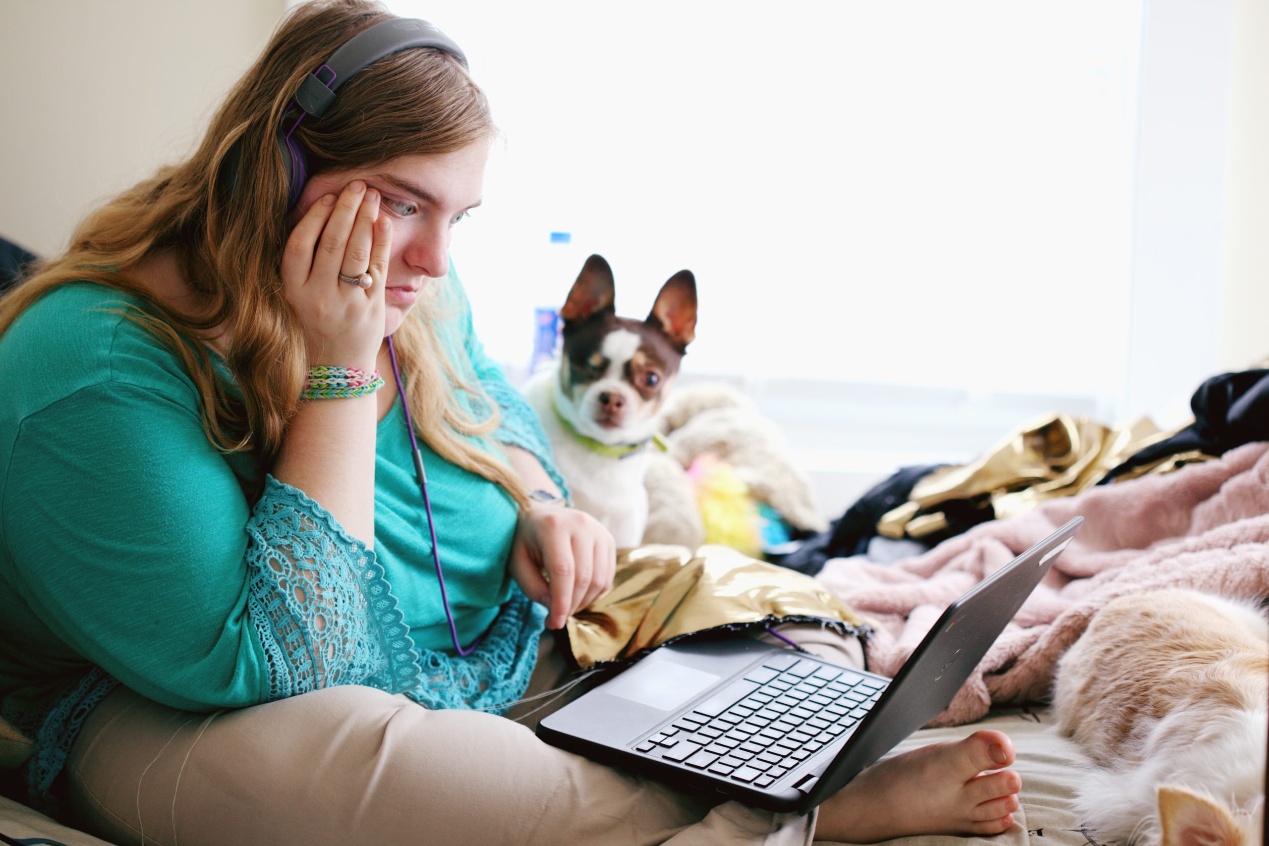 A person in a teal shirt and khakis sits on a bed while looking at a computer. A dog is sitting beside them.