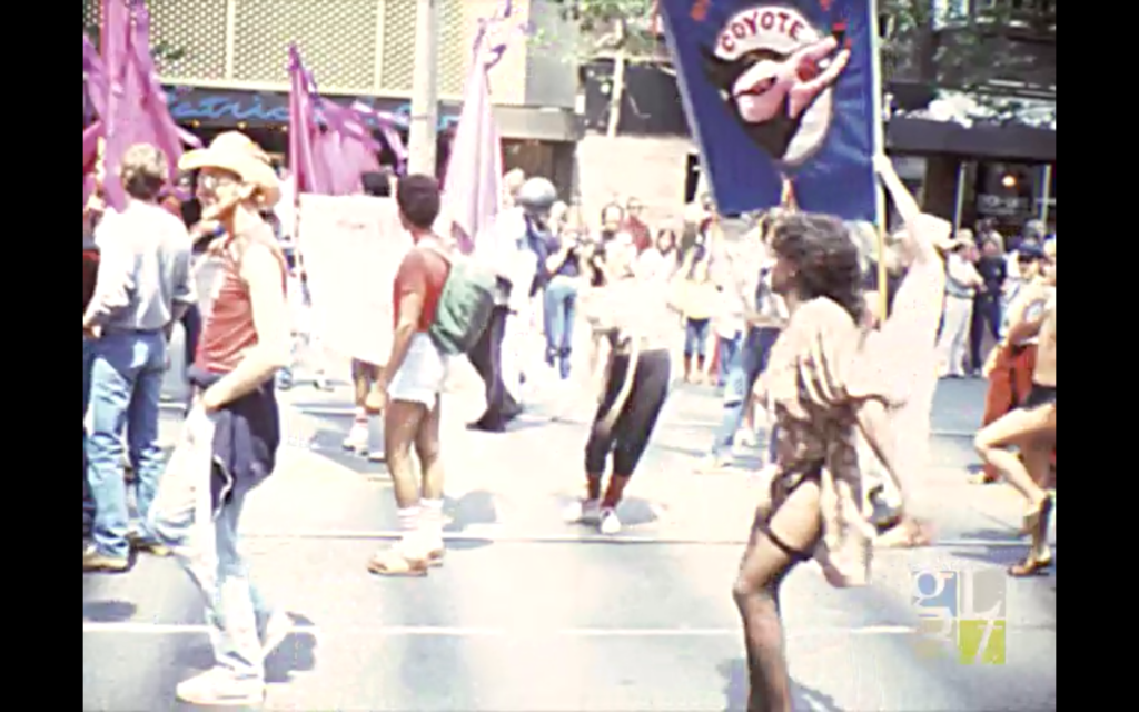 A still frame of aged VHS footage of people marching down a paved rod in the sunshine waving flags; one person in the foreground wears thigh-highs and carries a blue flag that reads COYOTE.